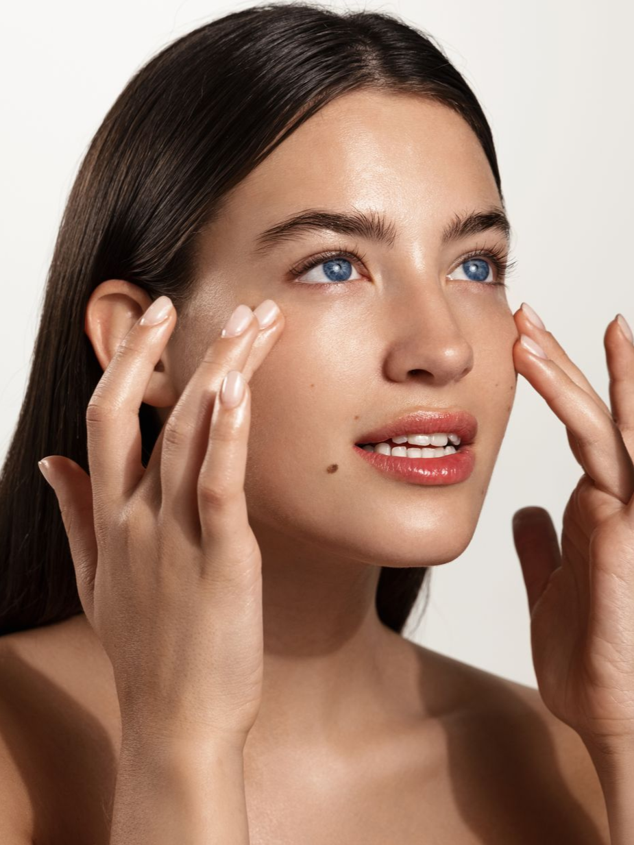 Woman applying skincare cream to her cheeks against a plain white background