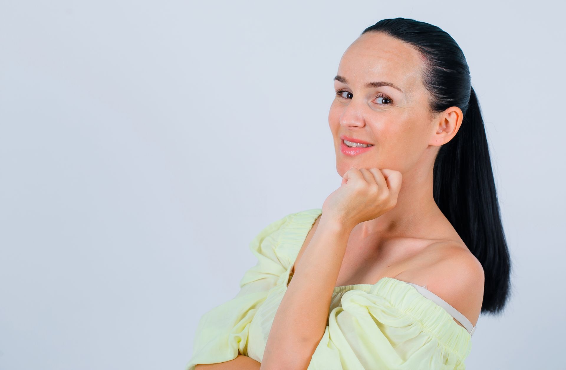 Woman with dark hair smiling in a light yellow off-shoulder top against a plain gray background