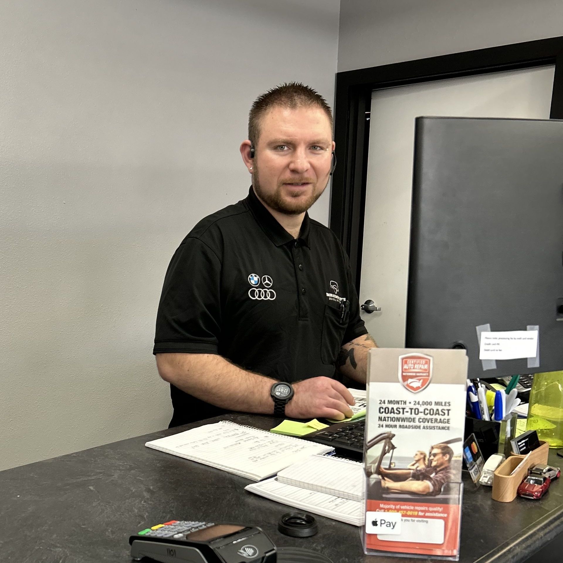Man at counter in black polo shirt, working at a car service desk.