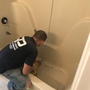 A man in a black shirt and jeans drills into the base of an off-white bathtub, potentially repairing or installing something.