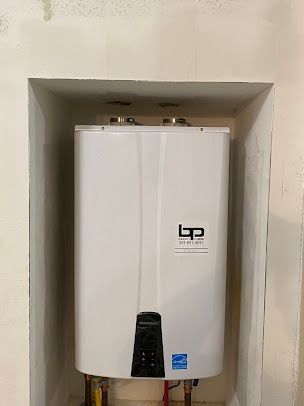 White tankless water heater installed in a recessed wall space. It has a black control panel and metal exhaust pipes on top.