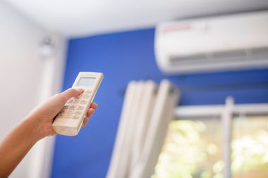Hand holding a remote pointed at a wall-mounted air conditioner in a blue-walled room