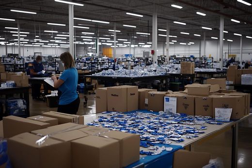 Busy warehouse with rows of boxes and workers packing items at long tables