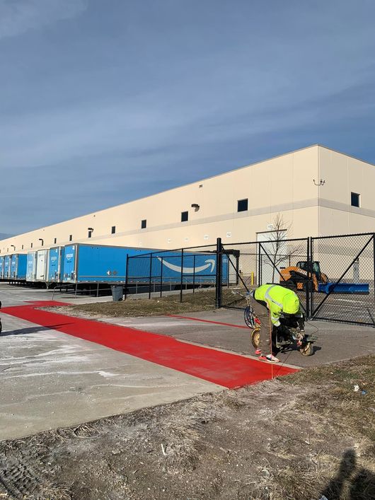 Worker painting a red curb line beside a beige warehouse with blue construction trailers.