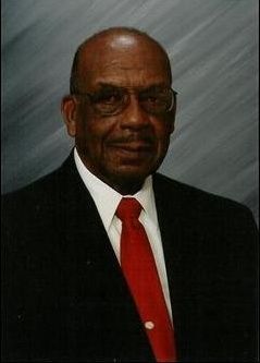 Man in black suit and red tie, wearing glasses, smiling slightly.