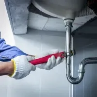 Gloved worker tightening a chrome sink drain pipe under a bathroom sink with a red wrench