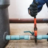 Gloved worker using a pipe cutter on a blue horizontal pipe with a valve in an industrial setting