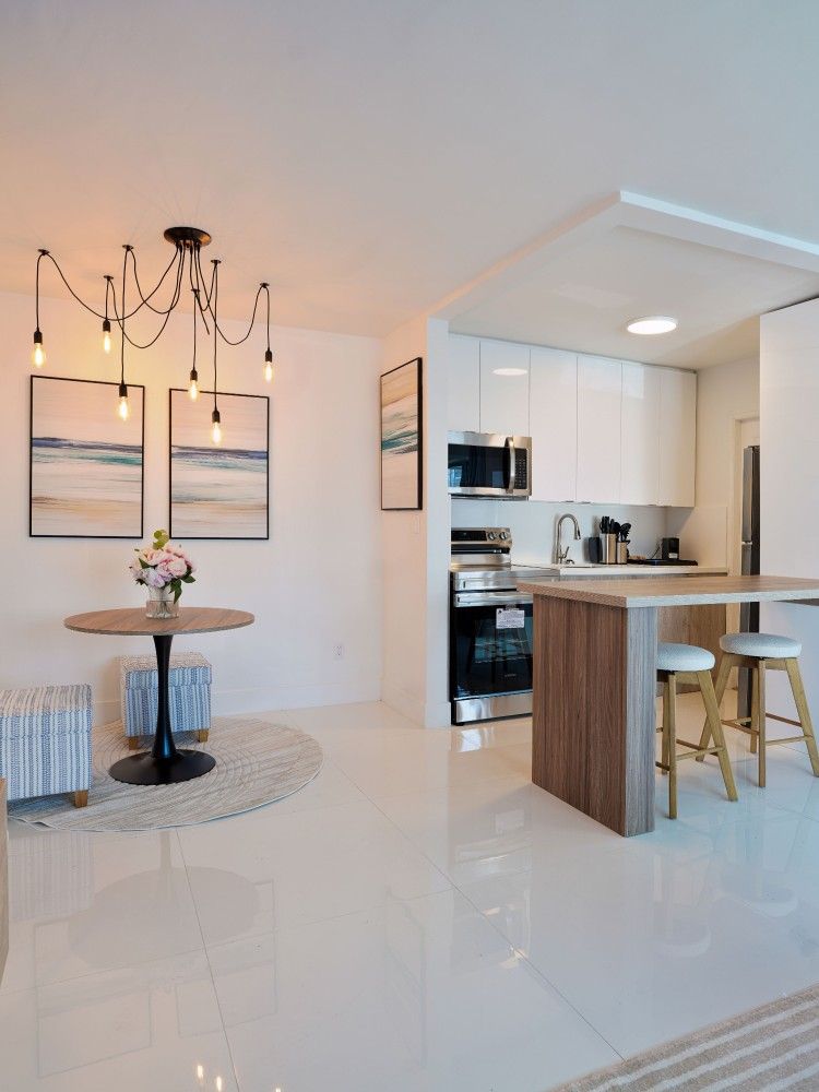 Bright modern kitchen and dining area with white tile floors, wood island, and round table near abstract wall art