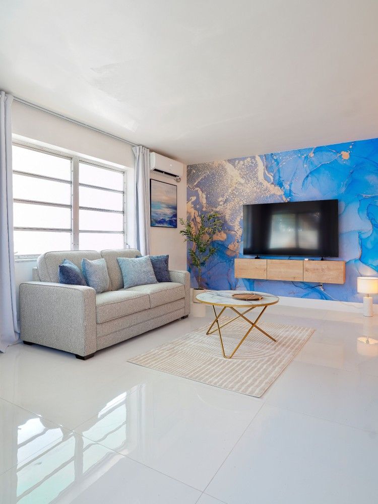 Modern living room with light gray sofa, wall-mounted TV, blue floral accent wall, and gold coffee table.