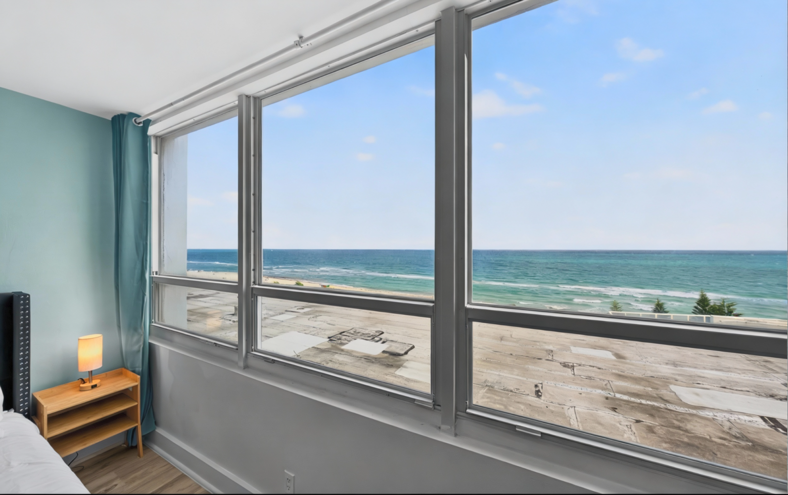 Bedroom window overlooking a sandy beach and ocean, with a small bedside lamp and side table.