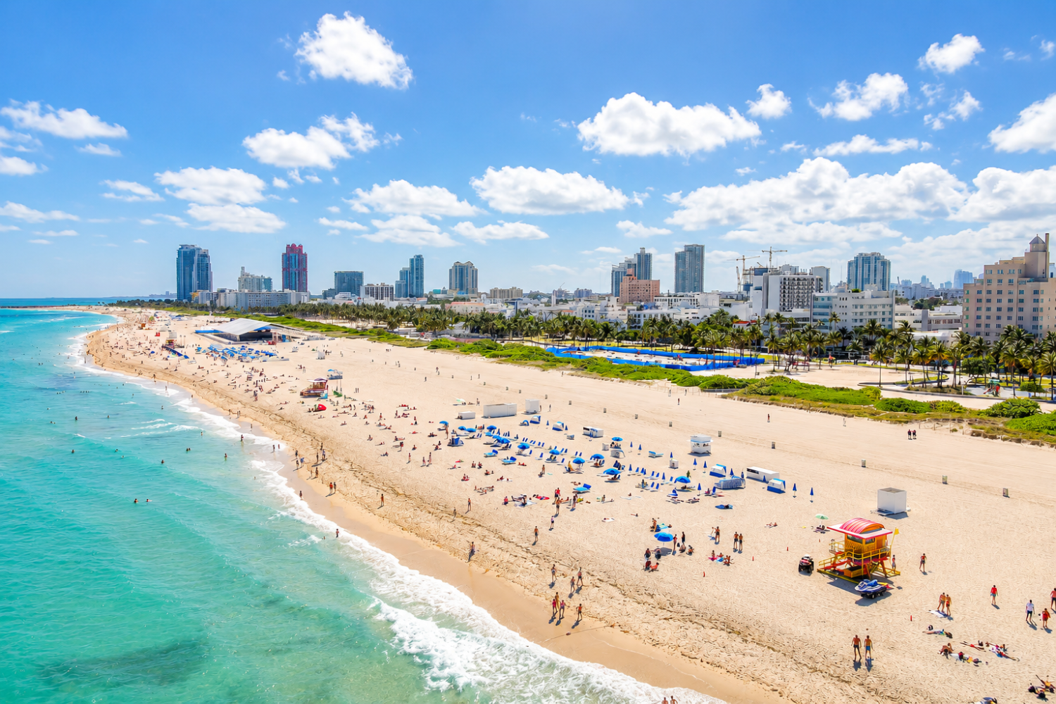 Sunny beach with turquoise water, crowds of beachgoers, and a city skyline under a blue sky