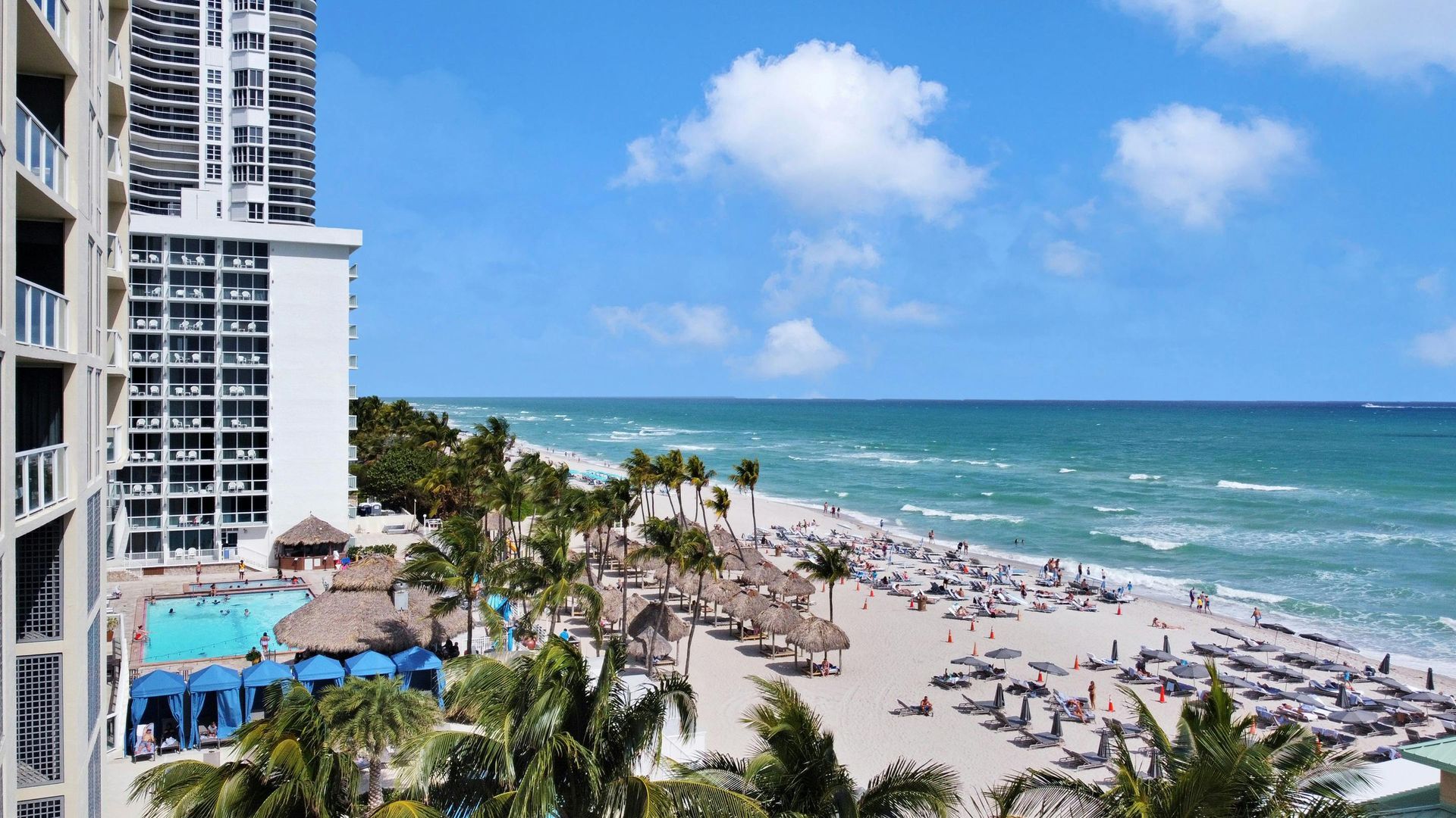 Beachfront resort with high-rise buildings, palm trees, lounge chairs, and turquoise ocean under a blue sky