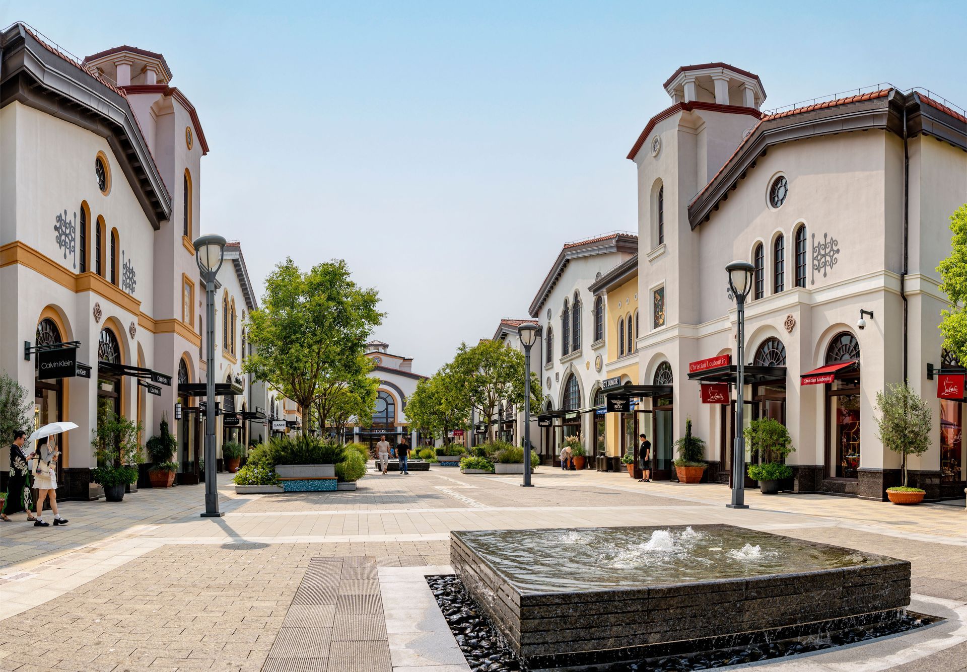 Bright shopping promenade with white stucco buildings, arched windows, trees, and a central fountain