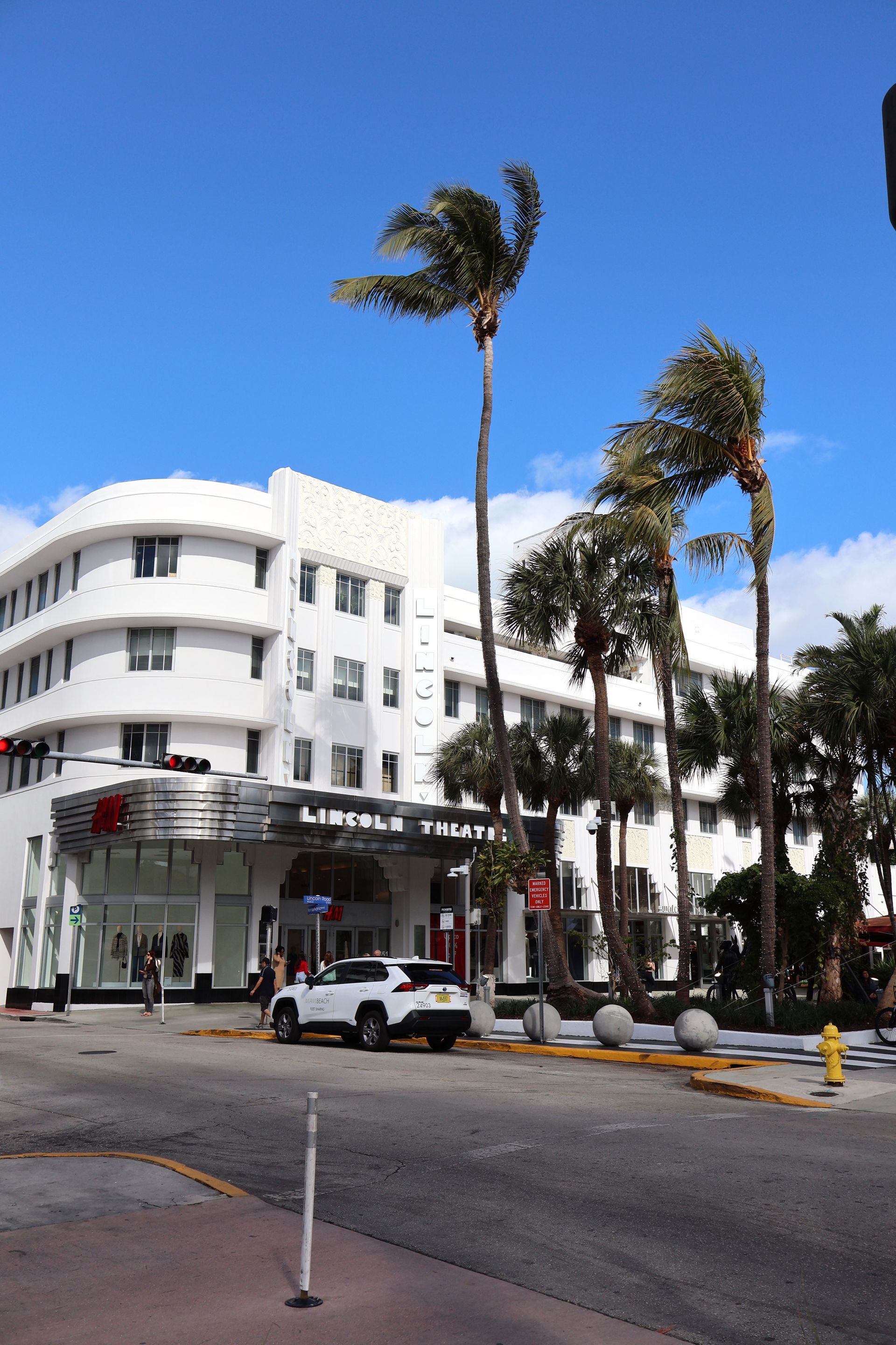 White Art Deco-style hotel at a sunny street corner with palm trees and parked cars