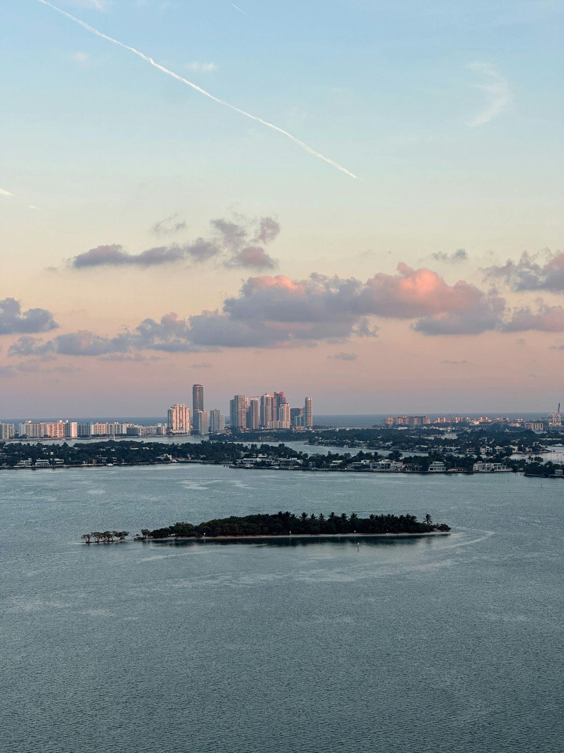 City skyline at dusk beyond a calm bay, with small island in the foreground and pink clouds overhead