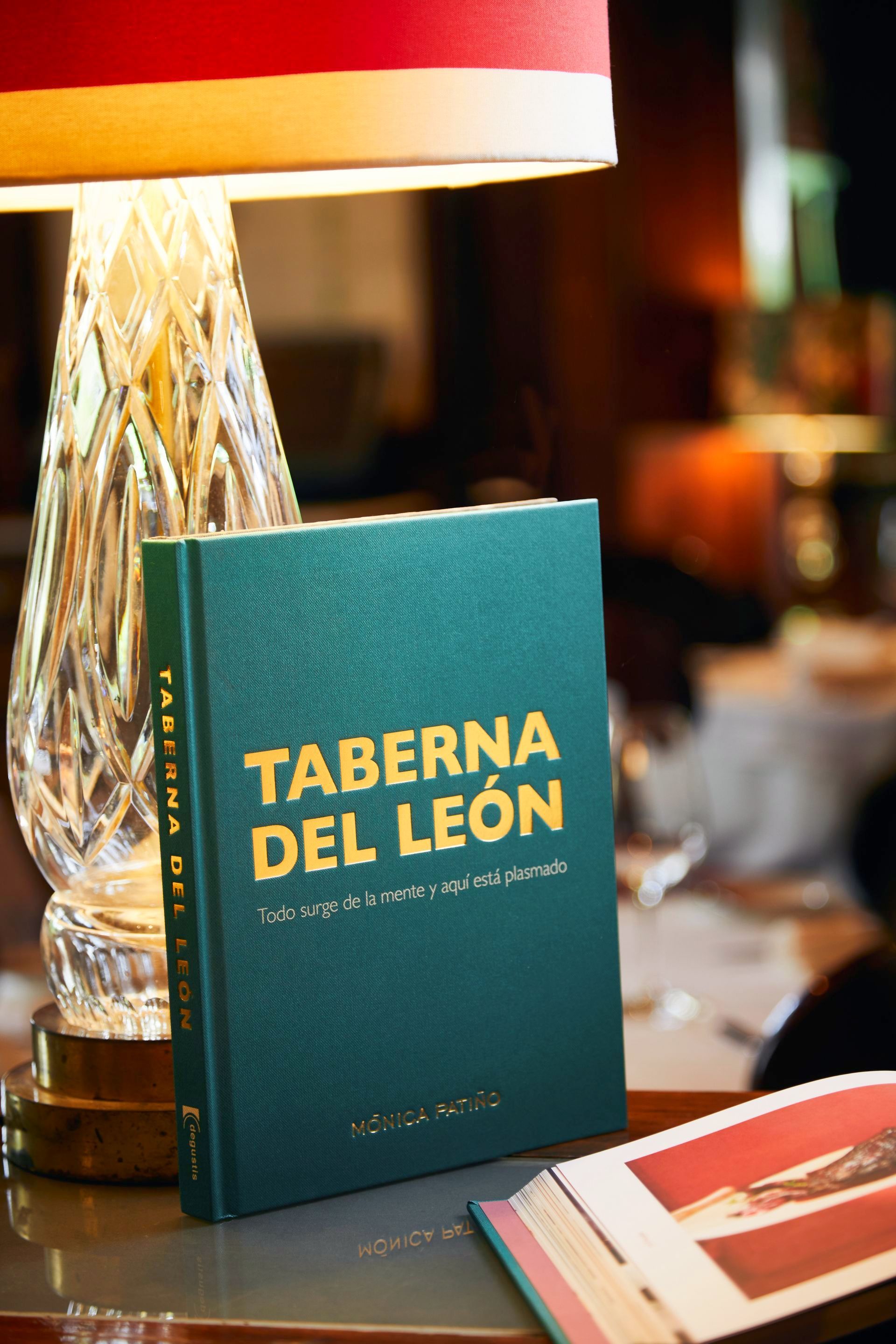 Menú color turquesa de “TABERNA DEL LEÓN” junto a una lámpara sobre una mesa, con un restaurante desenfocado al fondo.