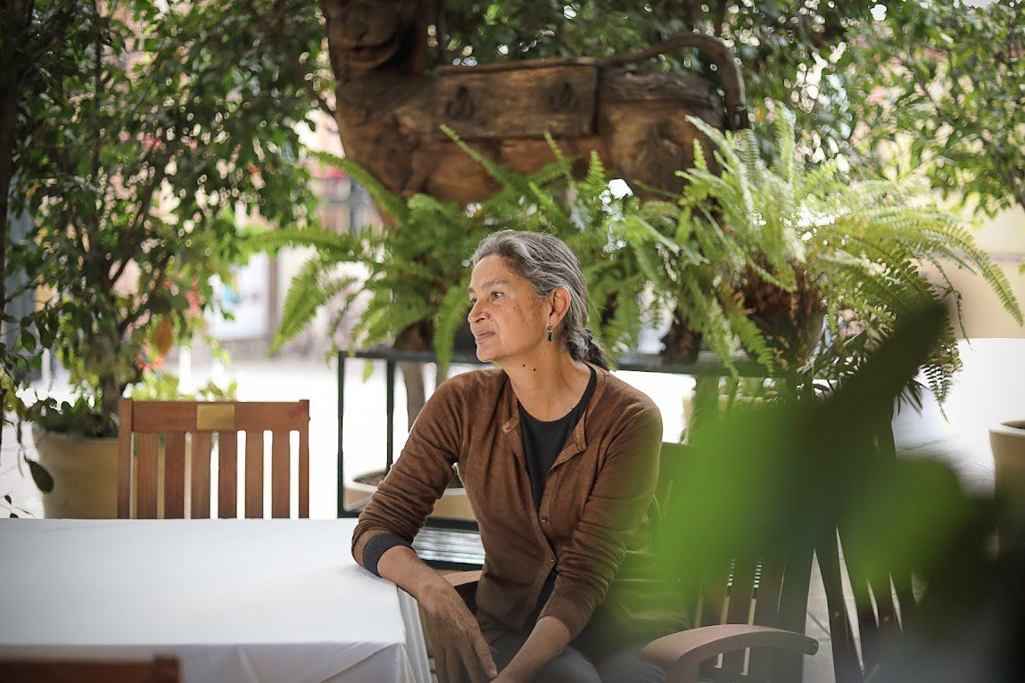 Una persona sentada en una mesa al aire libre bajo un árbol, mirando hacia la izquierda, con vegetación en primer plano.