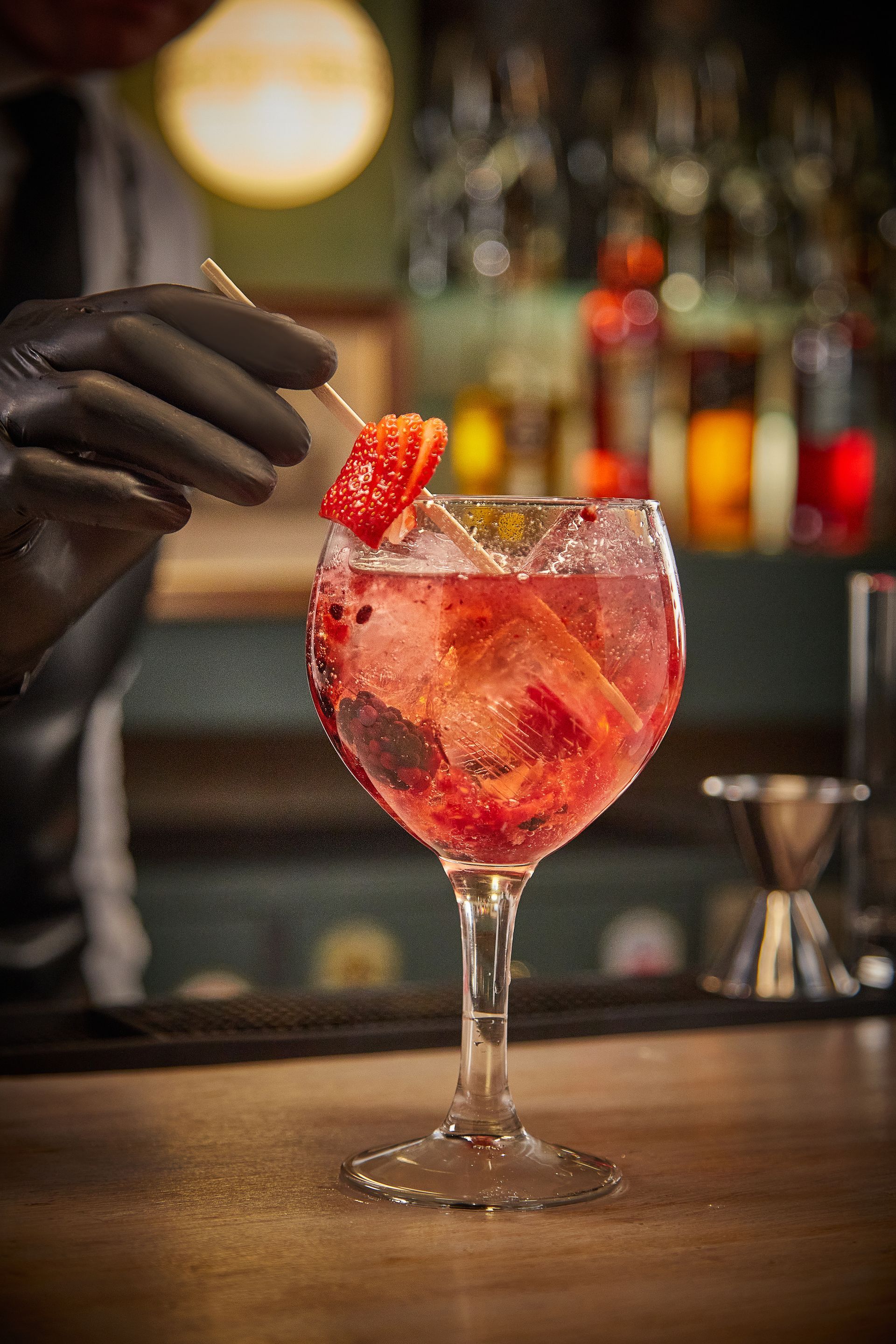 Un camarero con guantes decora un cóctel rojo con una fresa en un bar con poca luz.