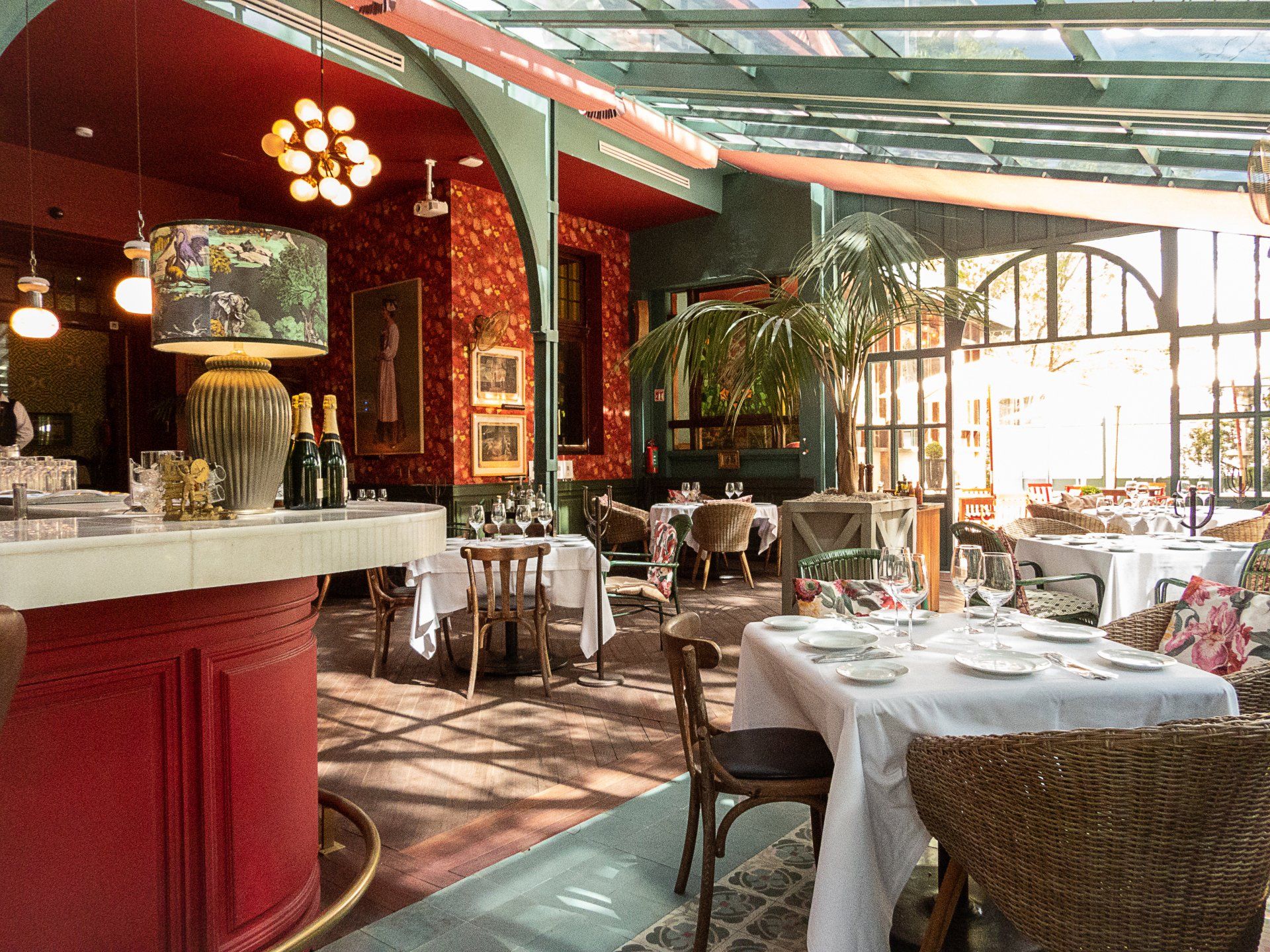 Restaurante luminoso y soleado con barra roja, manteles blancos y sillas de mimbre junto a grandes ventanales.