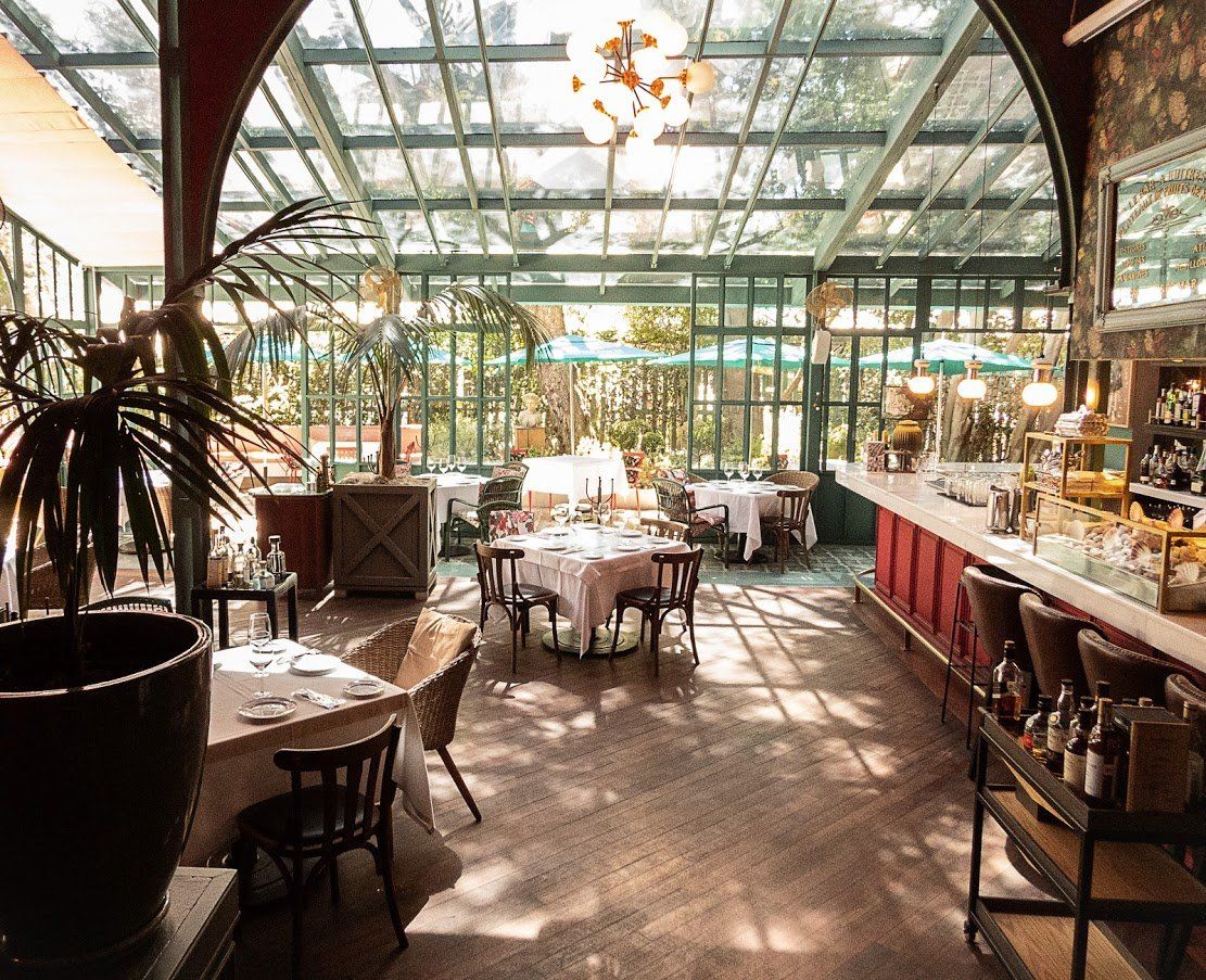 Cafetería luminosa con luz natural, tipo invernadero, con mesas, plantas y barra roja.