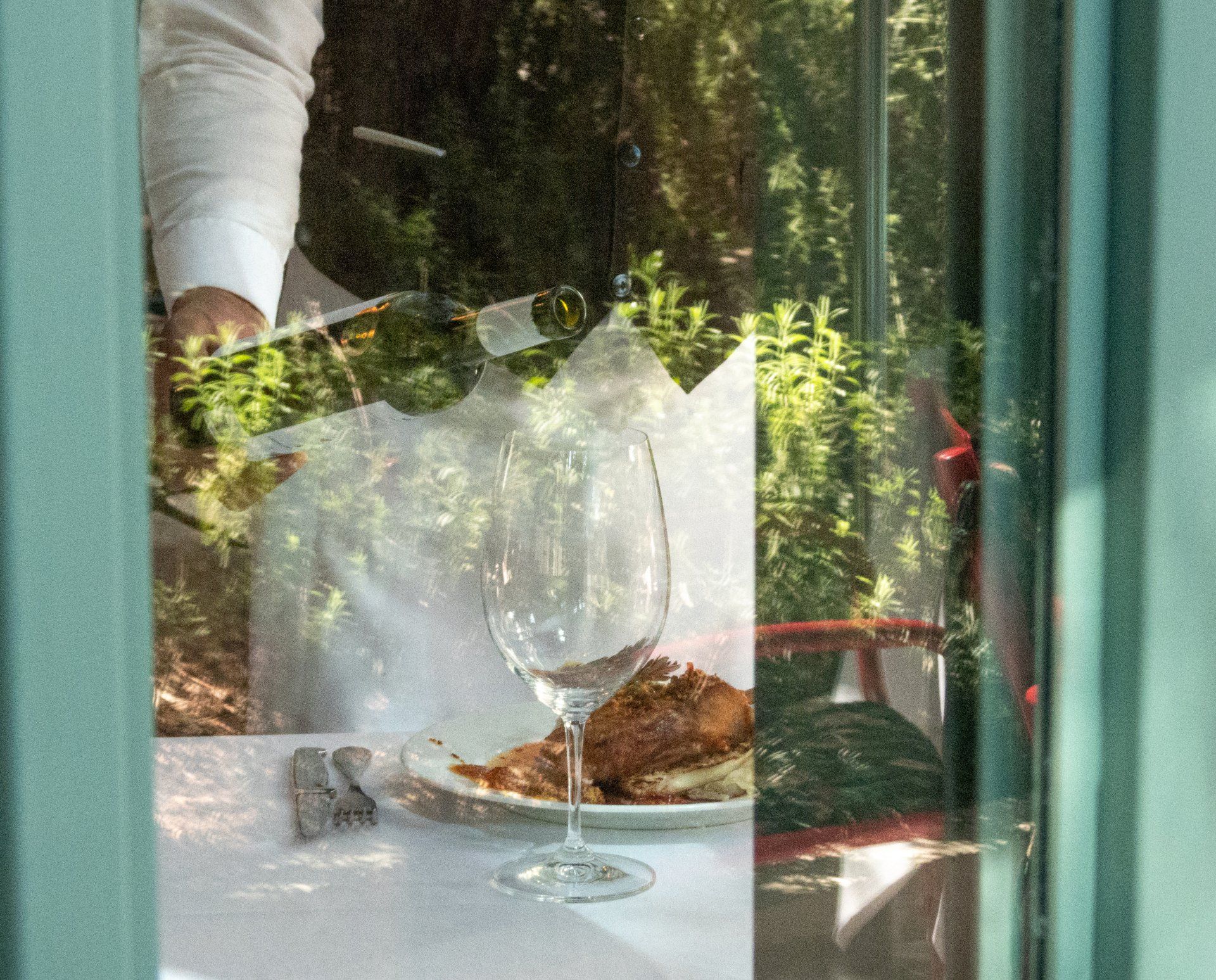 Mesa puesta junto a una ventana con copas de vino, pan y plantas que se reflejan en el cristal.