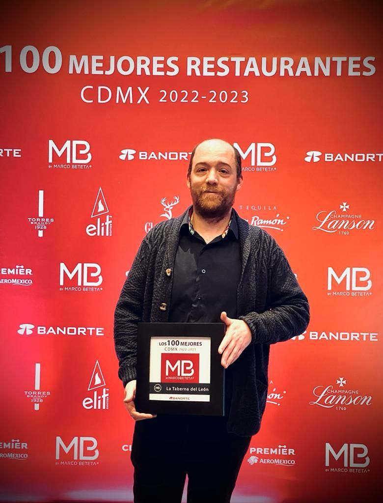 Man holding a plaque at a red awards backdrop reading “100 Mejores Restaurantes CDMX 2022-2023.”