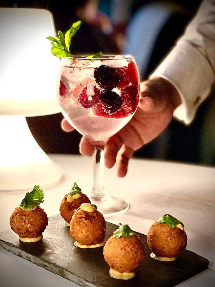 Una mano sostiene un cóctel rosa con frutos rojos y menta junto a aperitivos del tamaño de un bocado en una bandeja oscura.