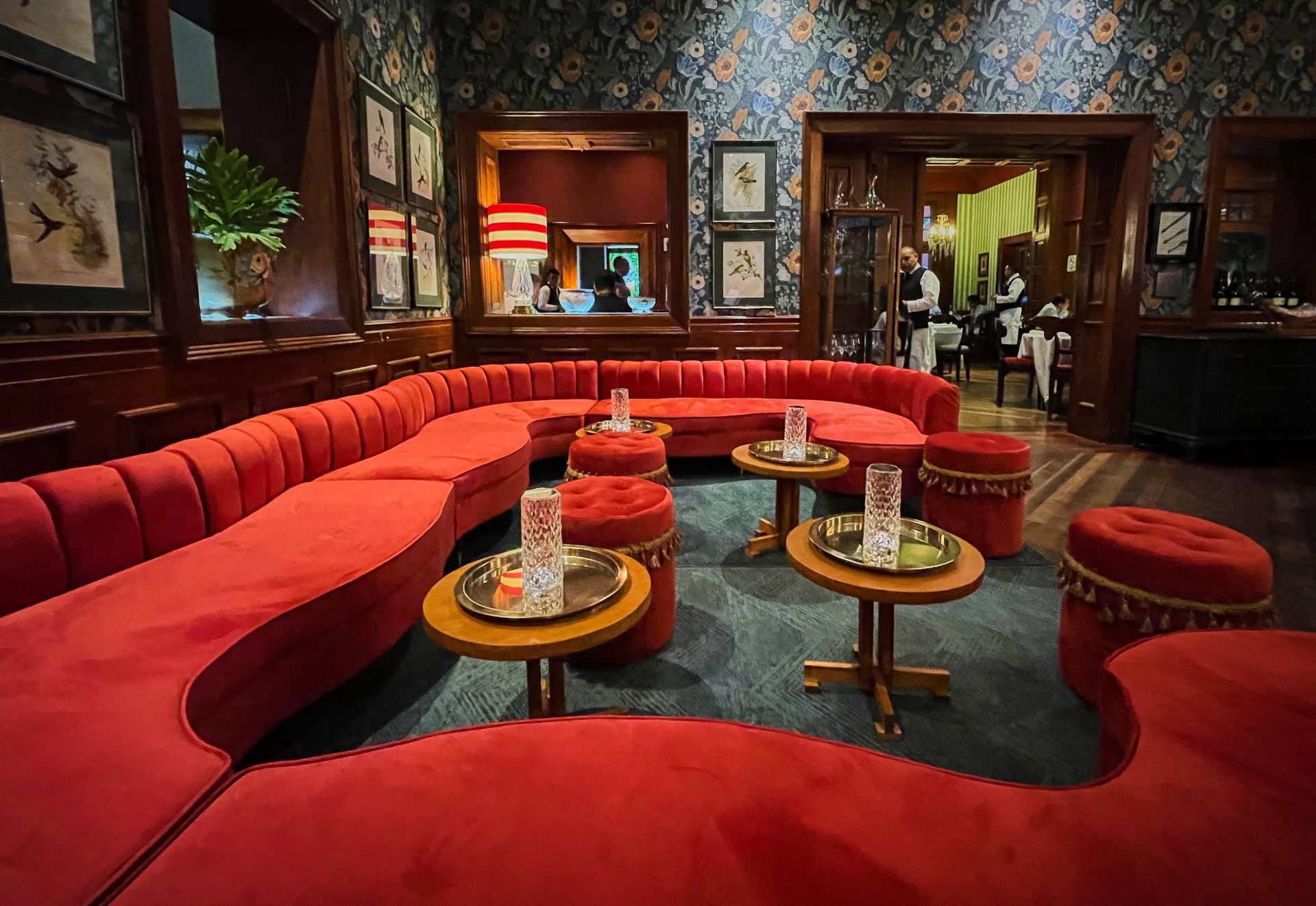 Salón de terciopelo rojo con sofás curvos, mesas redondas y papel pintado estampado en un bar con iluminación tenue.