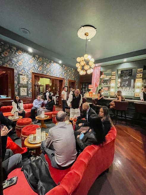 Gente sentada en un acogedor salón de café con asientos de terciopelo rojo, lámparas de araña y papel tapiz colorido.