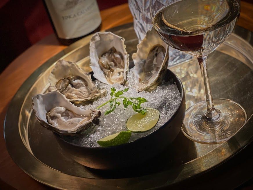 Plato de ostras sobre hielo con rodajas de lima y una copa de vino tinto en una bandeja.