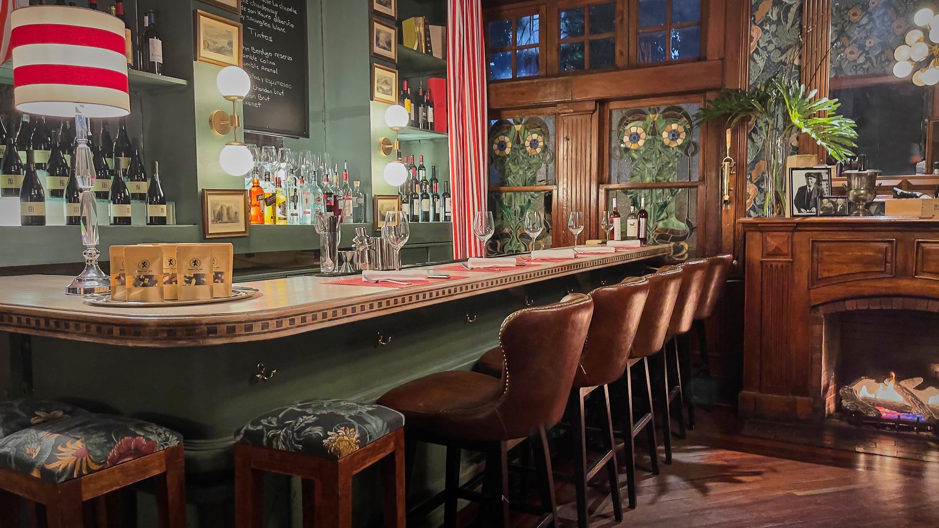 Interior de bar acogedor con una larga barra, taburetes marrones, iluminación cálida y estantes con botellas y decoración.