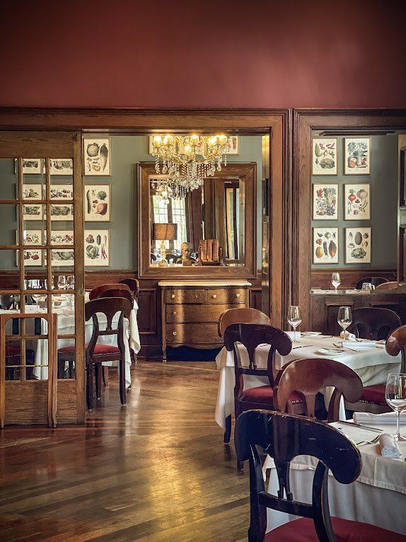Elegante comedor con lámpara de araña, mesas y sillas de madera, suelo pulido y cuadros enmarcados en paredes rojas.