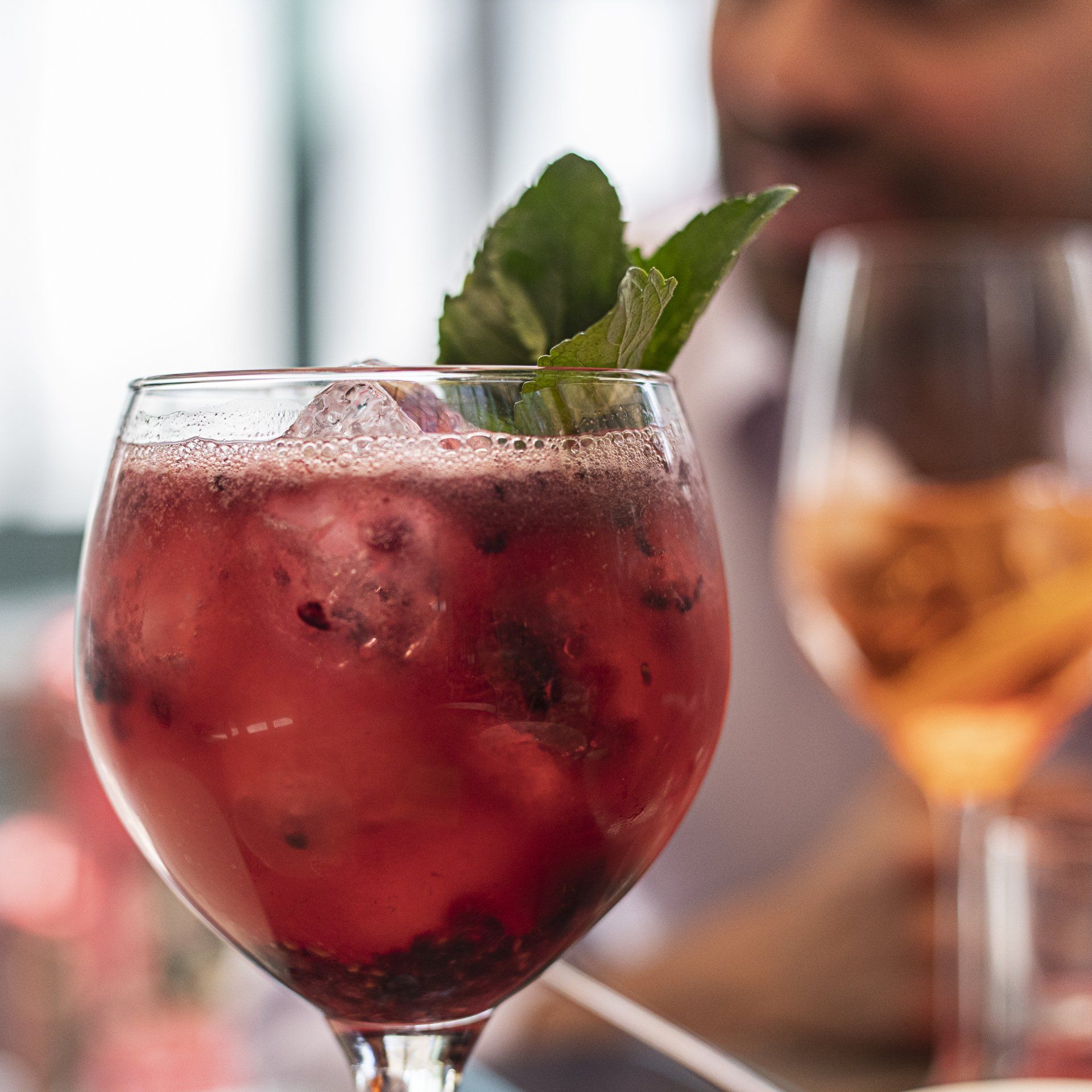 Cóctel de frutos rojos con guarnición de menta en una copa de tallo, con la bebida y la persona borrosas al fondo.