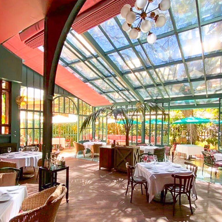 Cafetería al aire libre bañada por el sol, con mesas con manteles blancos bajo un toldo de cristal y paredes rojas.