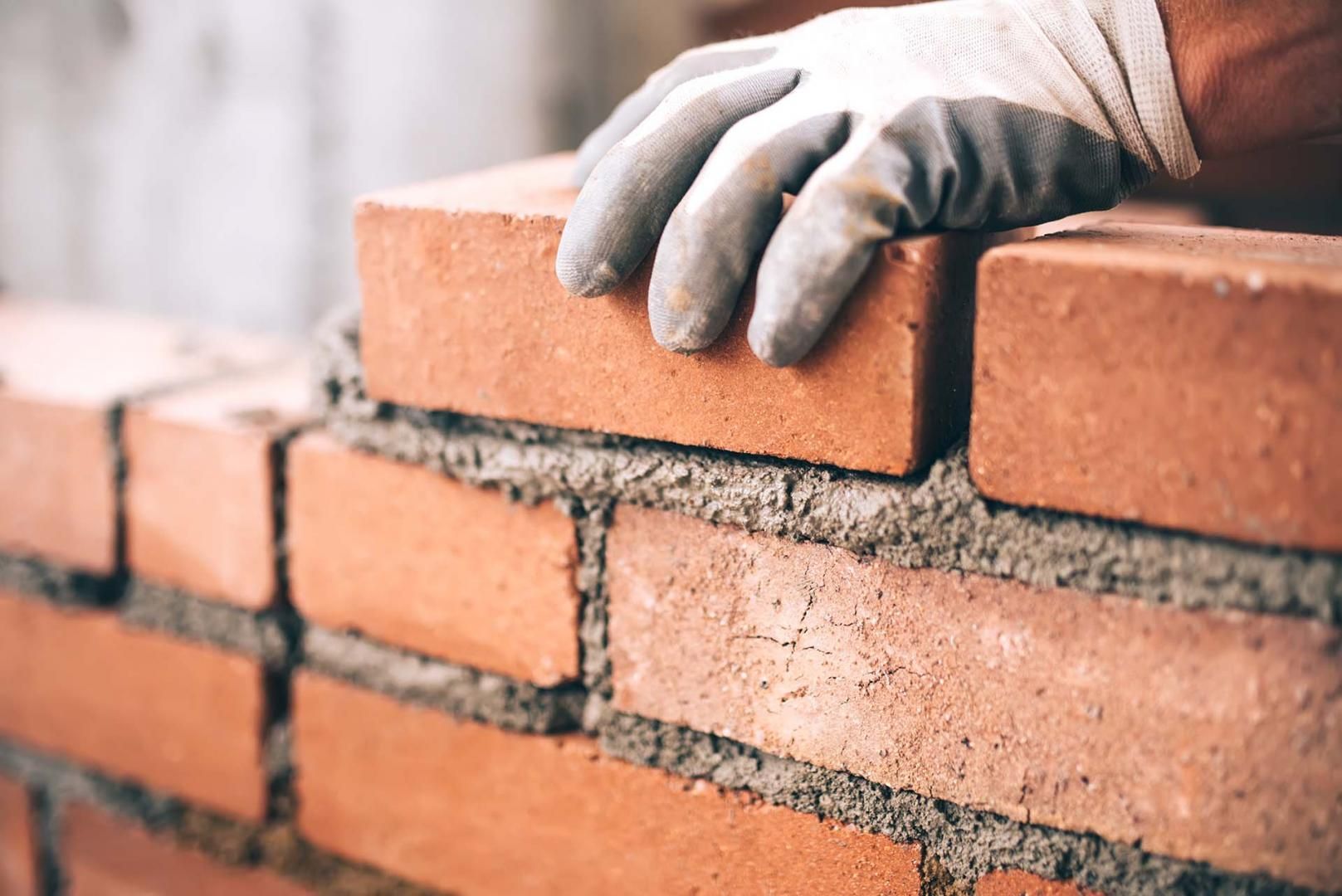 Main gantée posant une brique sur un mur de briques partiellement construit, à l'aide de mortier.