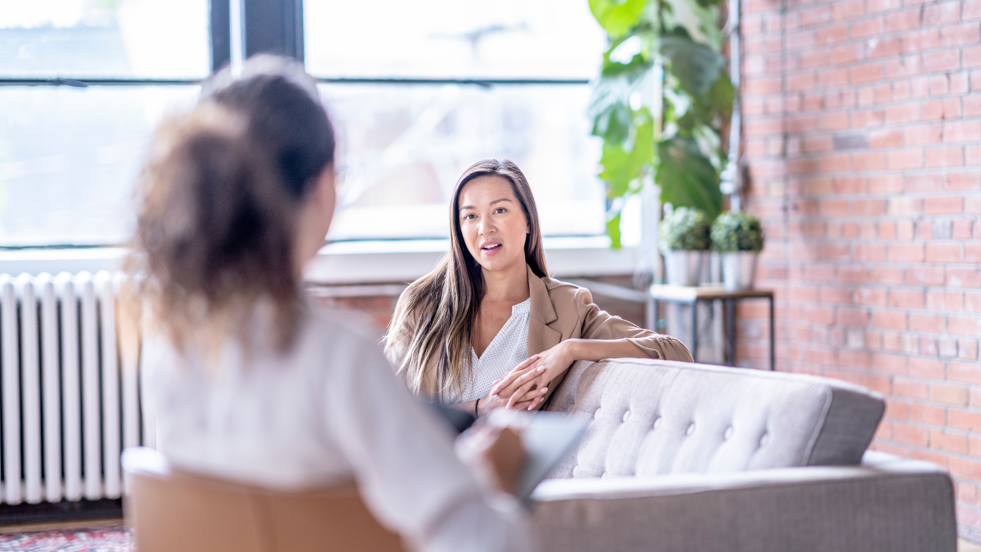 Woman therapist talking to a person seated in a chair, in a brick-walled office with a window and plants.