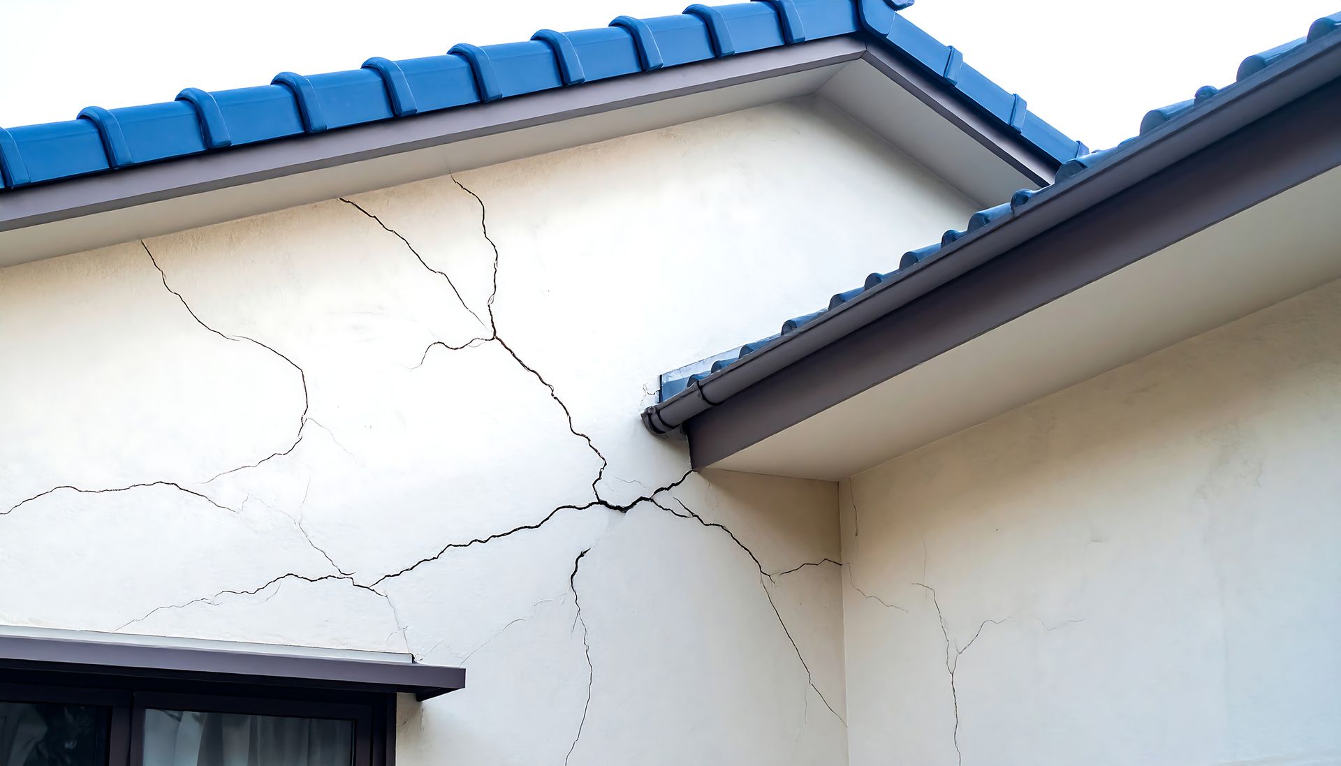 A exterior cream-colored house wall featuring prominent spiderweb-like cracks near the roofline and corner.