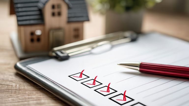 A red pen rests on a clipboard with a checked to-do list, positioned next to a miniature house model on a wooden table.