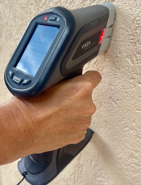 A hand-held lead paint analyzer pressed against a wall, displaying a digital reading screen with a red indicator light.