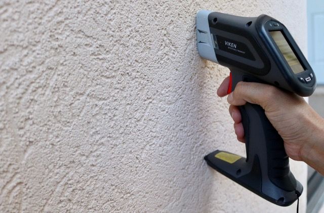 A hand holds a moisture meter against a baseboard near a wood-look floor, displaying data on a digital screen.