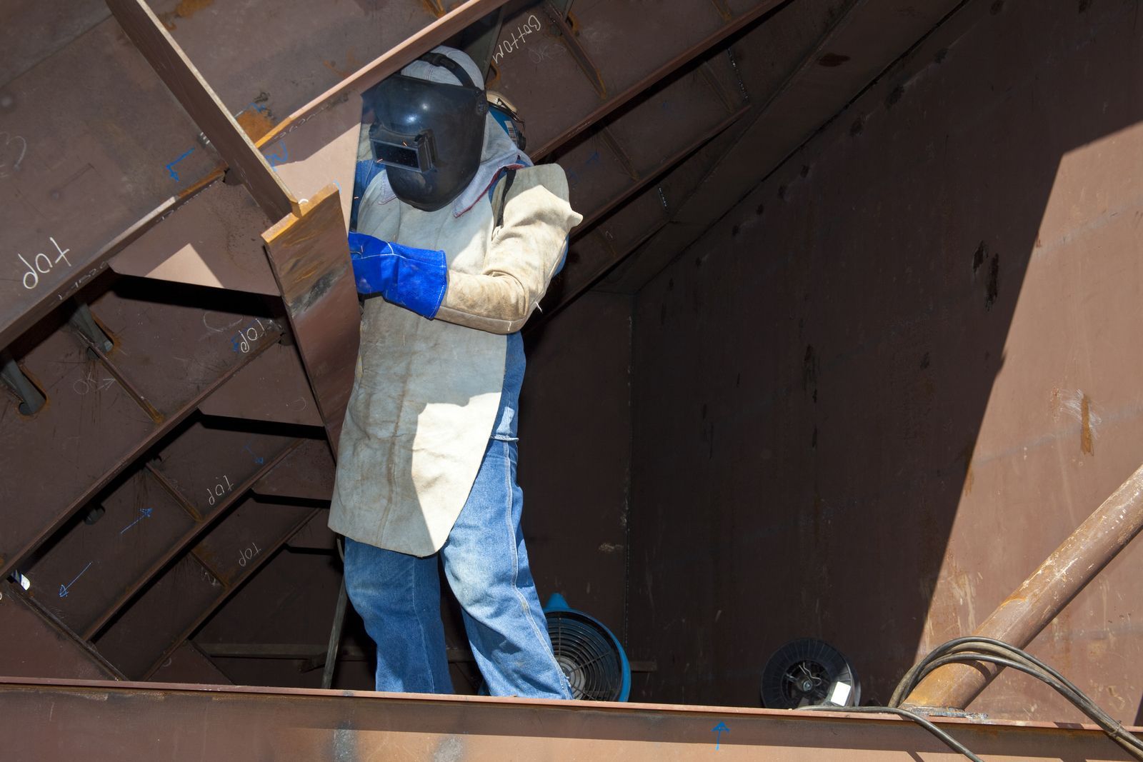 A welder in protective gear works inside a large metal structure, possibly the hull of a ship.