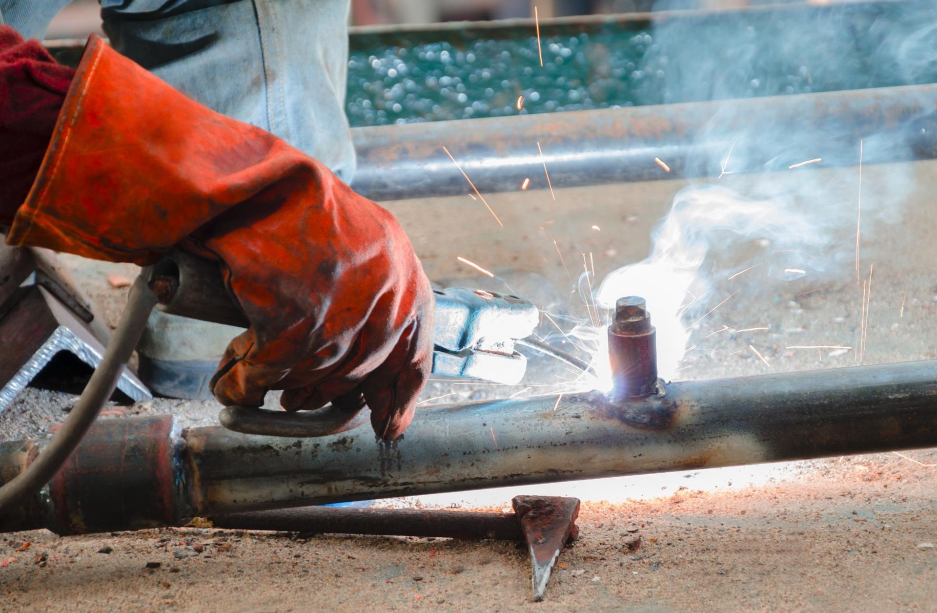 A welder wearing orange safety gloves fuses metal parts, creating bright sparks and smoke.