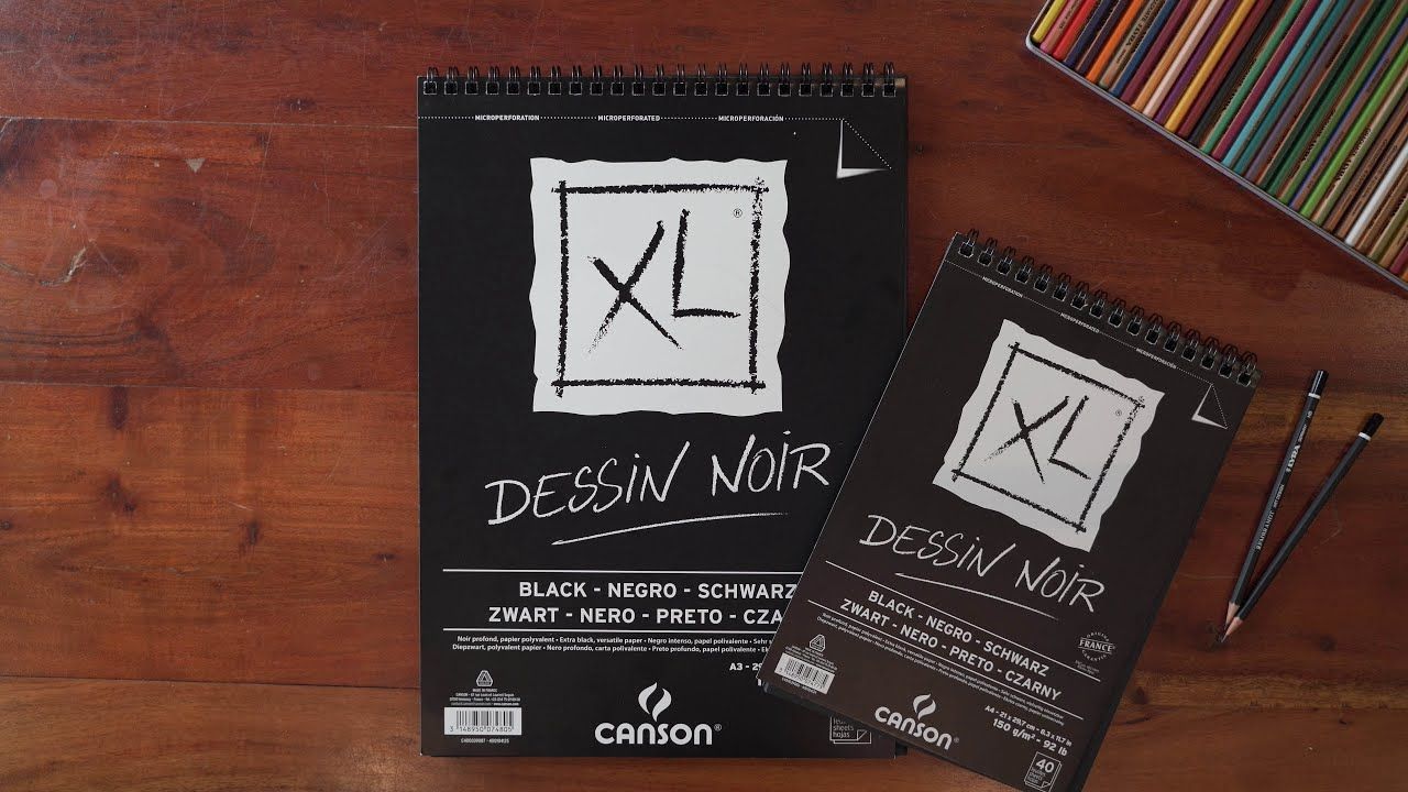 Canson XL Black paper 150gsm over a wodden table surrounded by colour pencils.