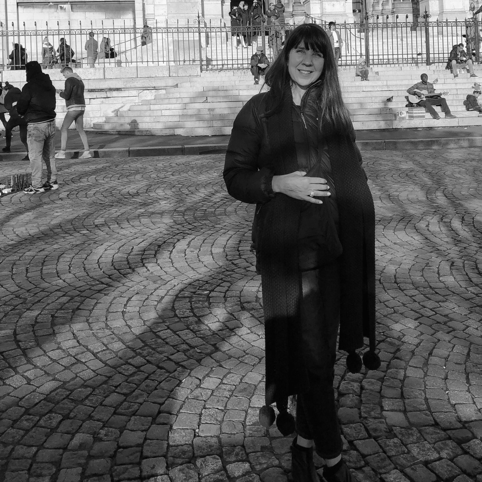 Paula Dakin standing on a cobblestone plaza, holding her stomach and smiling slightly.
