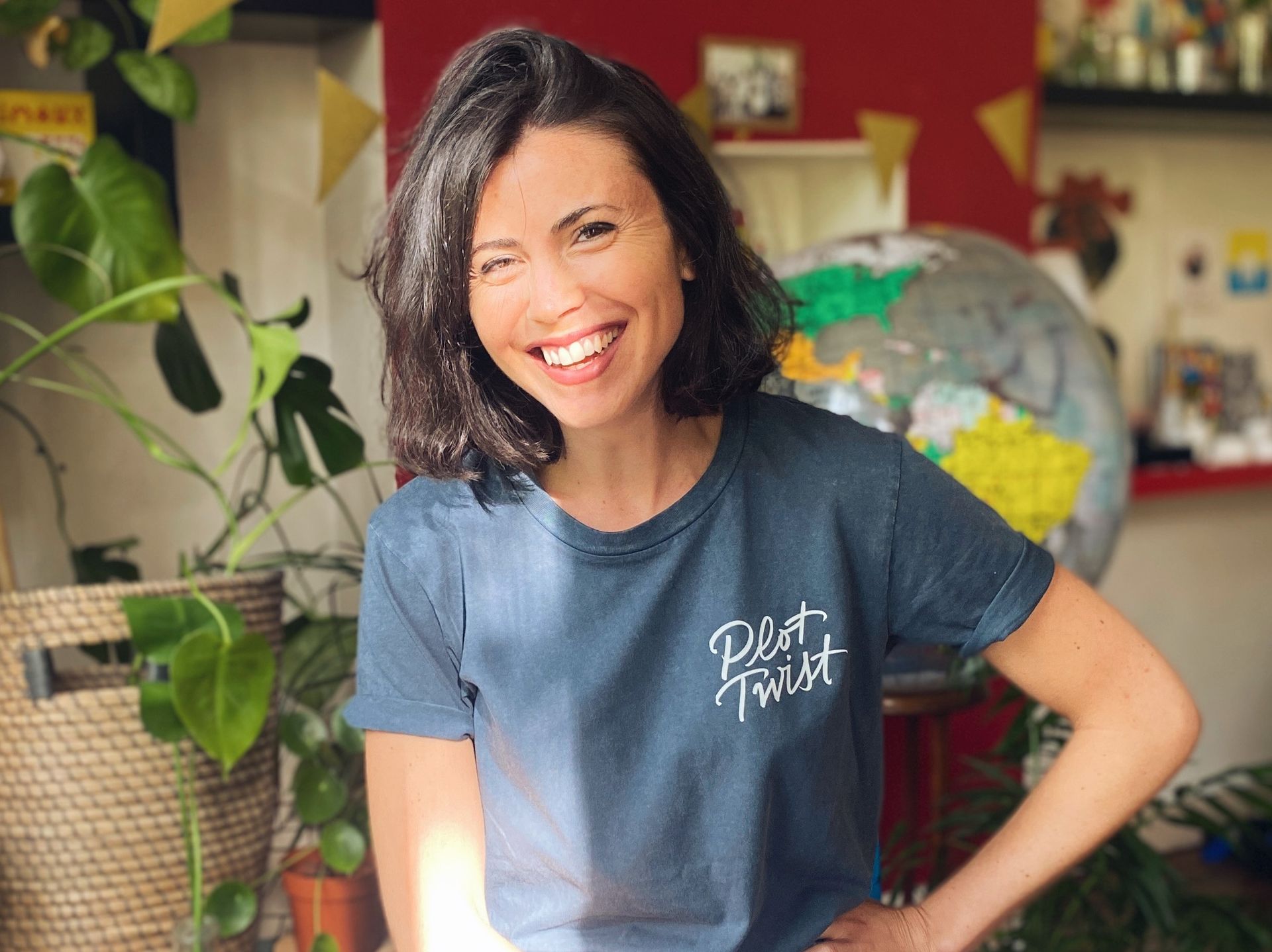 Smiling Gina Serret in a navy T-shirt standing indoors with plants and a globe in the background
