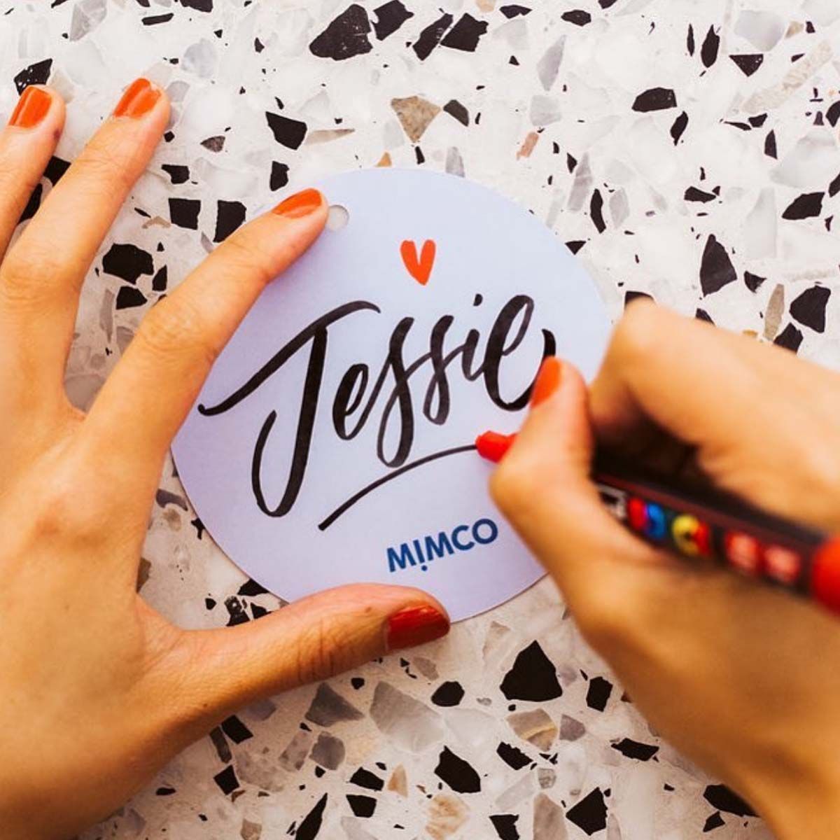Hands writing “Jessie” on a white round label with an orange heart, on a speckled tabletop.