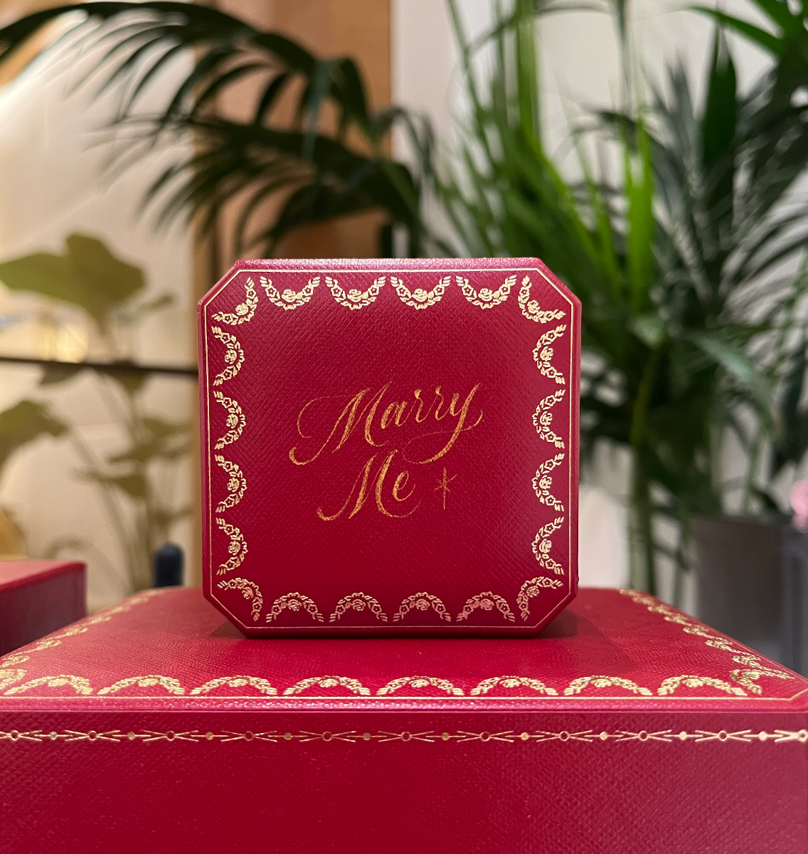 Red gift box with gold trim and “Merry Me” text, stacked on a larger red box indoors.