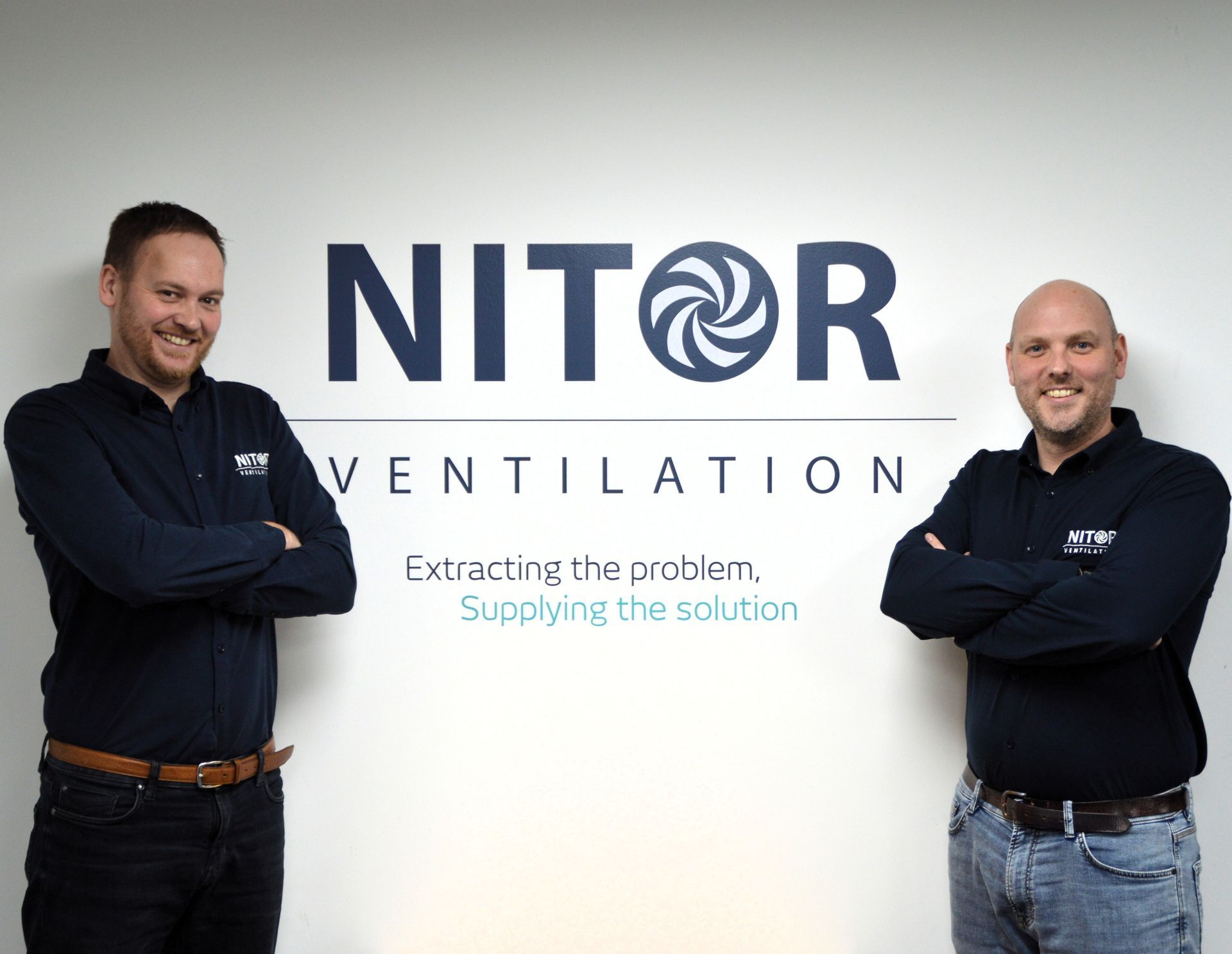 Two men in blue shirts stand in front of a white wall with the