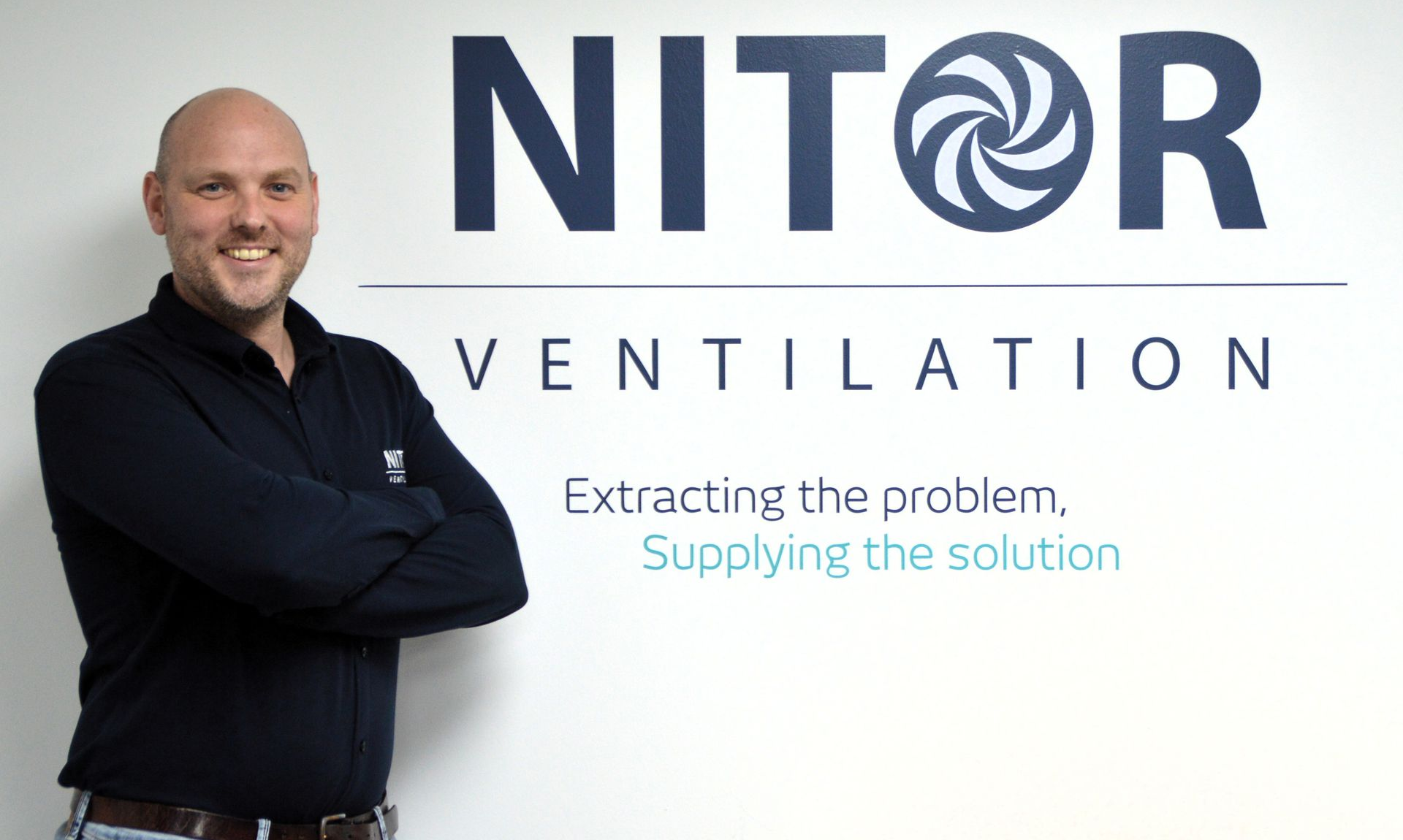 Man standing beside a sign for NITOR Ventilation. Black shirt, logo with a swirl, tagline: extracting the problem, supplying the solution.