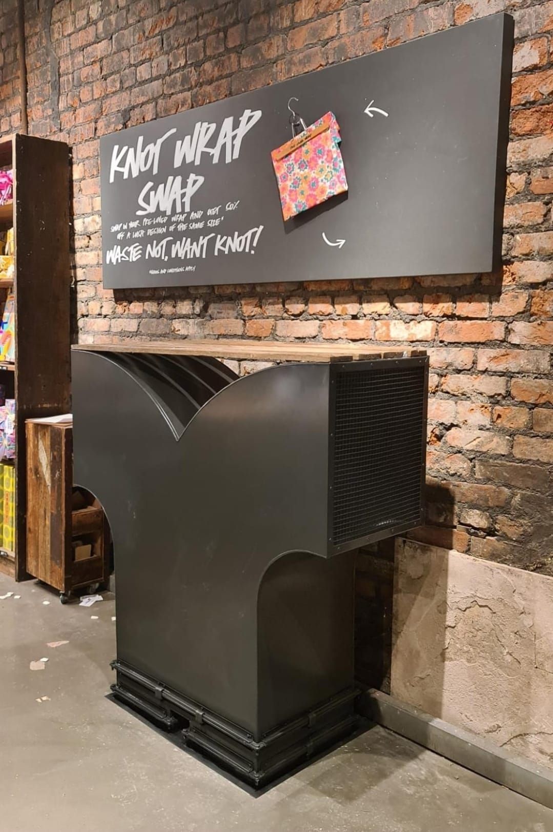 Black display kiosk against a brick wall with a chalkboard sign above it in a store or cafe.